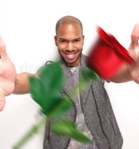 Grinning man in a grey vest shoving a blurry red rose at the camera
