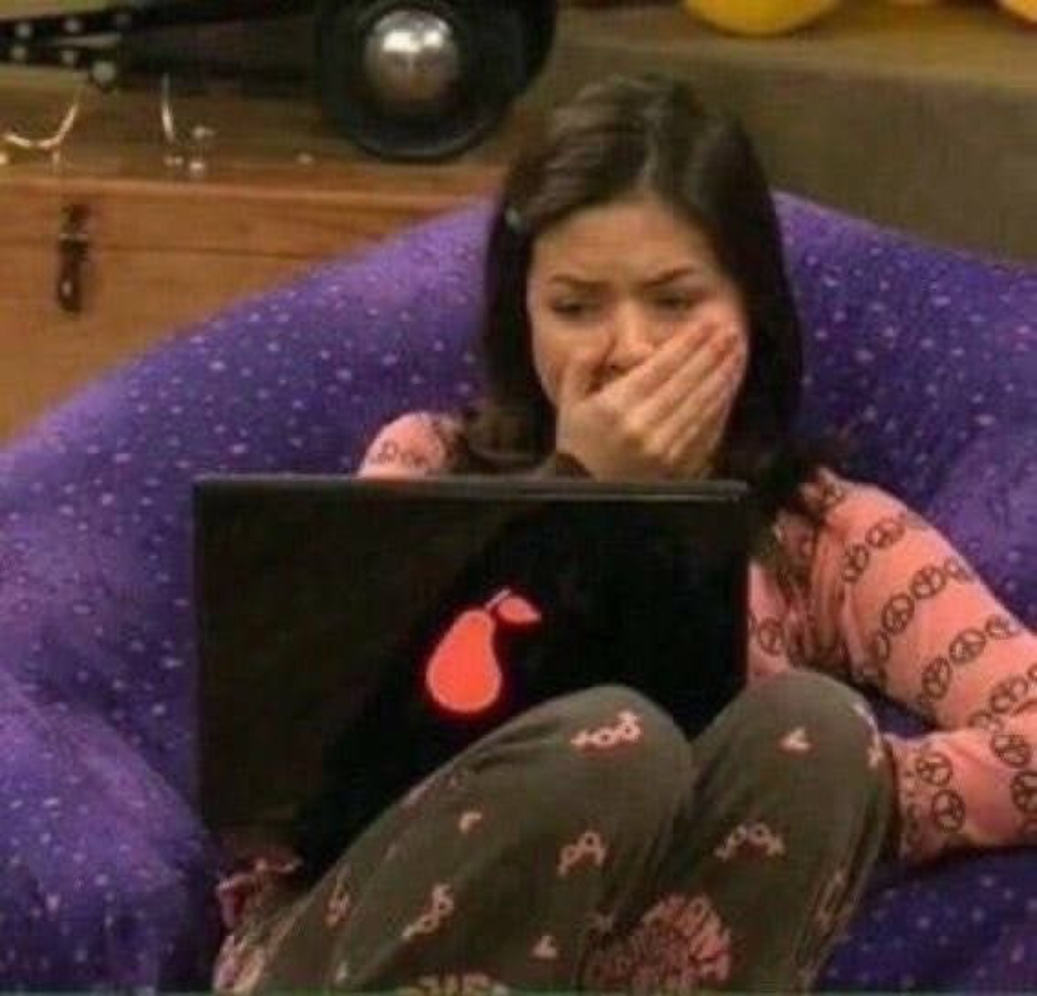 Girl in pajamas on a beanbag chair covering her mouth with her hand in shock while on her laptop