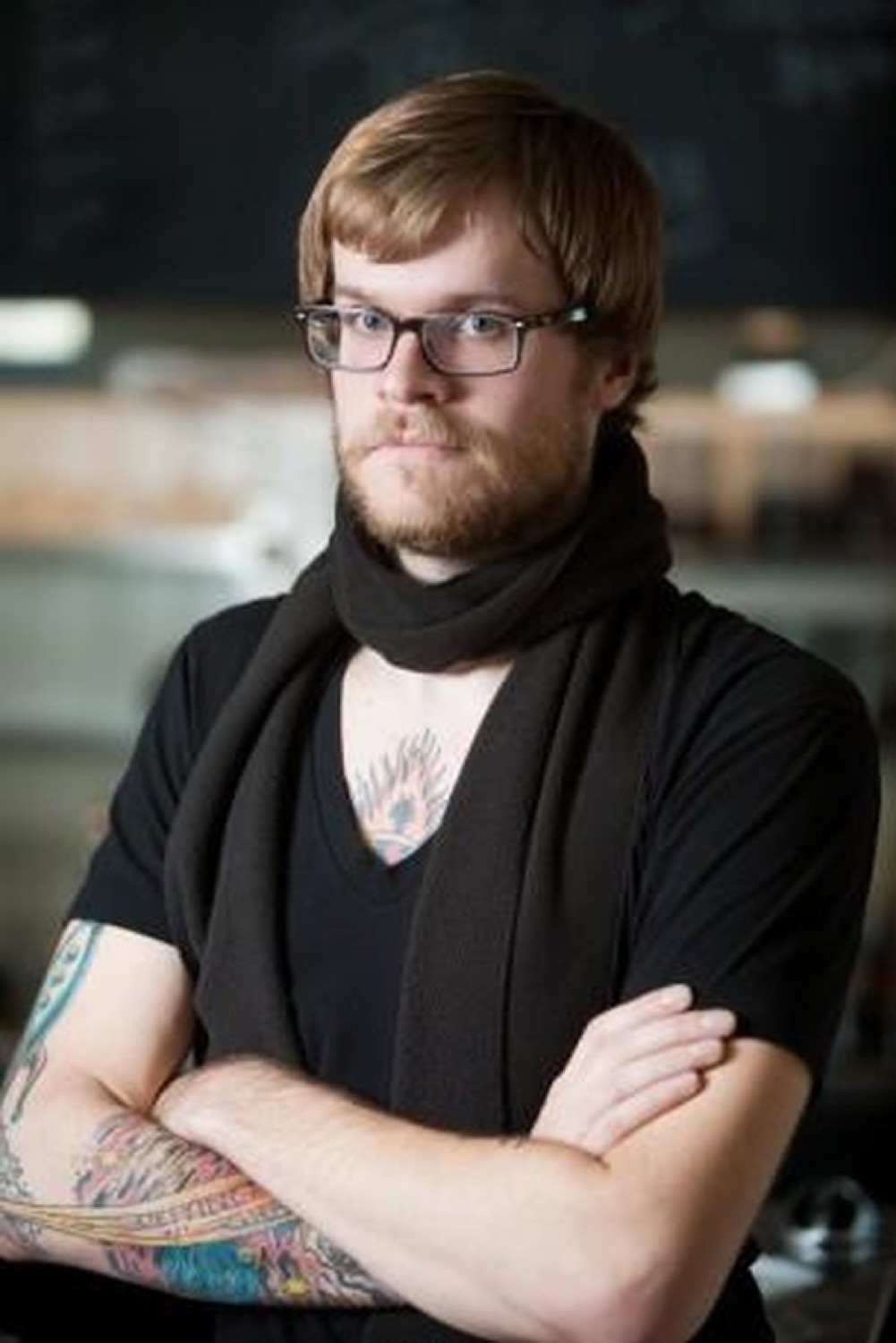 Bearded man in glasses and a scarf with arms crossed giving a flat, judgmental stare