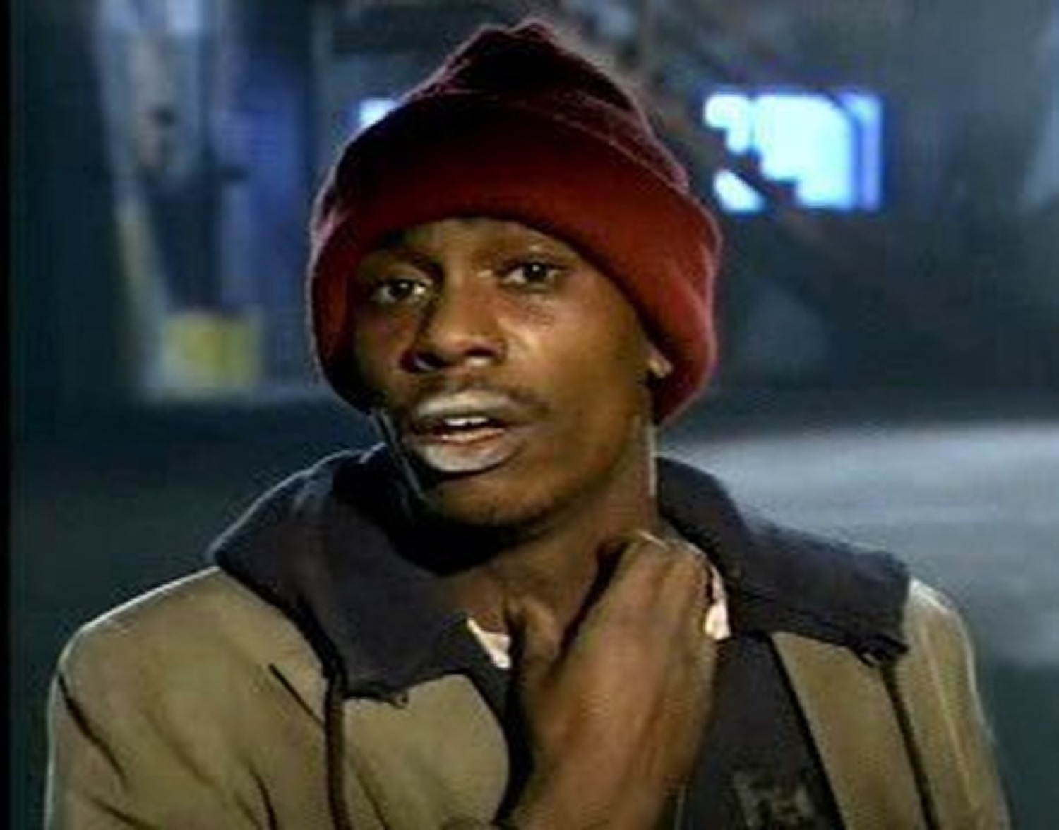 Dave Chappelle in a red beanie looking uneasy, hand at his throat like something's off