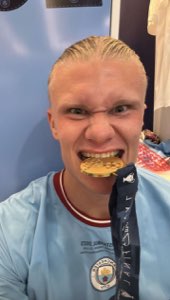Erling Haaland biting his Champions League medal with wild eyes and a massive grin
