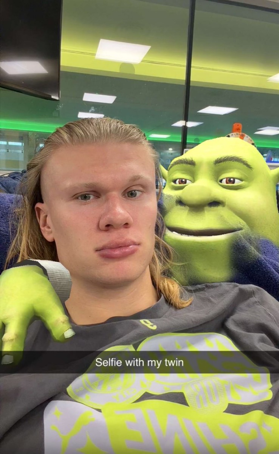 Blond footballer's deadpan selfie next to a grinning Shrek mascot, captioned 'Selfie with my twin'