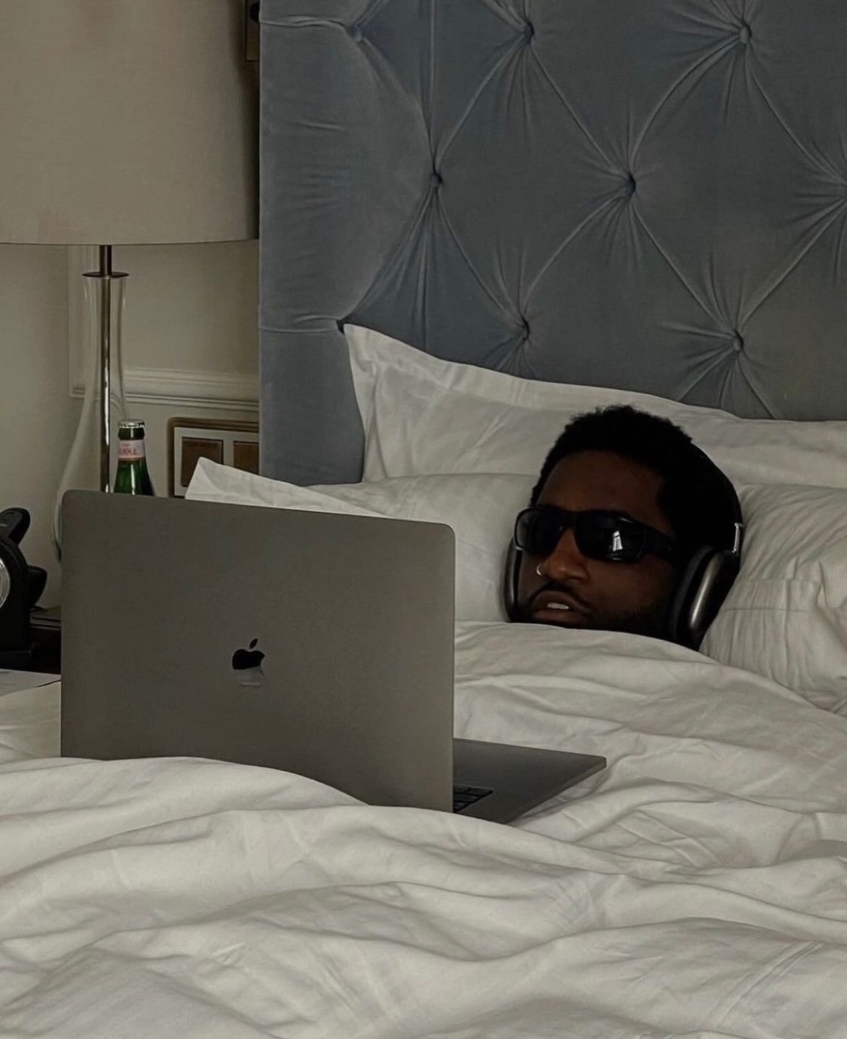 Man in sunglasses and headphones peeking from under the covers beside a laptop