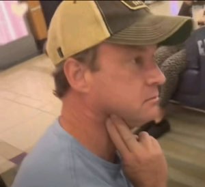 Man in a trucker hat holding his neck, staring off with a skeptical, deep-in-thought look