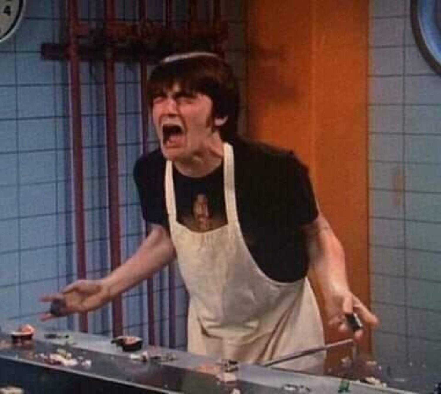 Guy in an apron behind a counter mid-scream, arms out in total meltdown