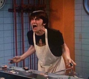 Guy in an apron behind a counter mid-scream, arms out in total meltdown
