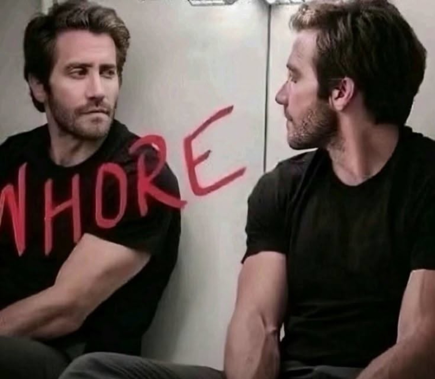 Two men sitting side by side, one glancing away with 'WHORE' scrawled in red across the wall and his shirt