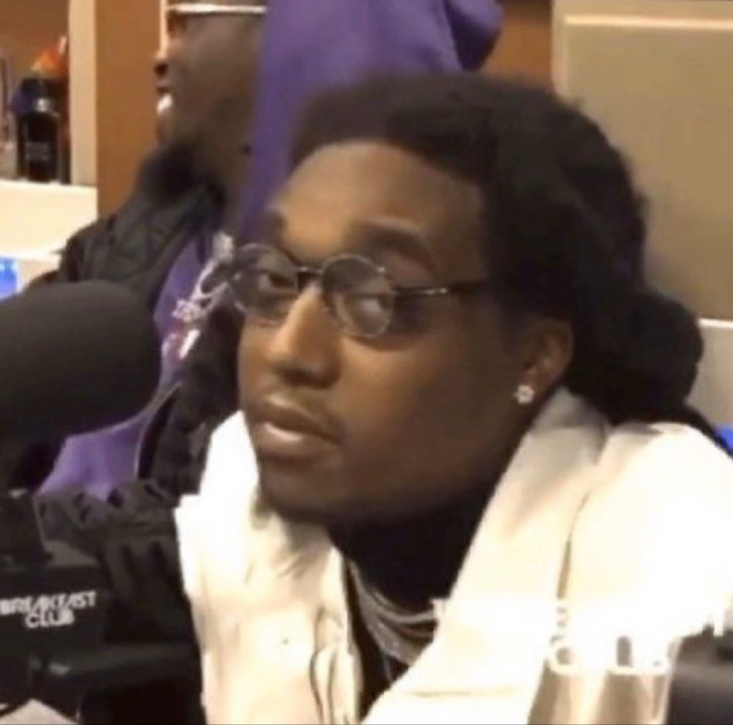 Man in round glasses at a radio mic giving a flat, unbothered side-eye