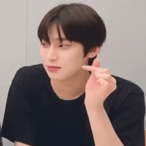 K-pop guy in a black tee doing a subtle finger heart with a soft smirk