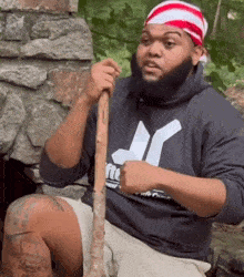 Man in a bandana and hoodie holding a stick and shrugging