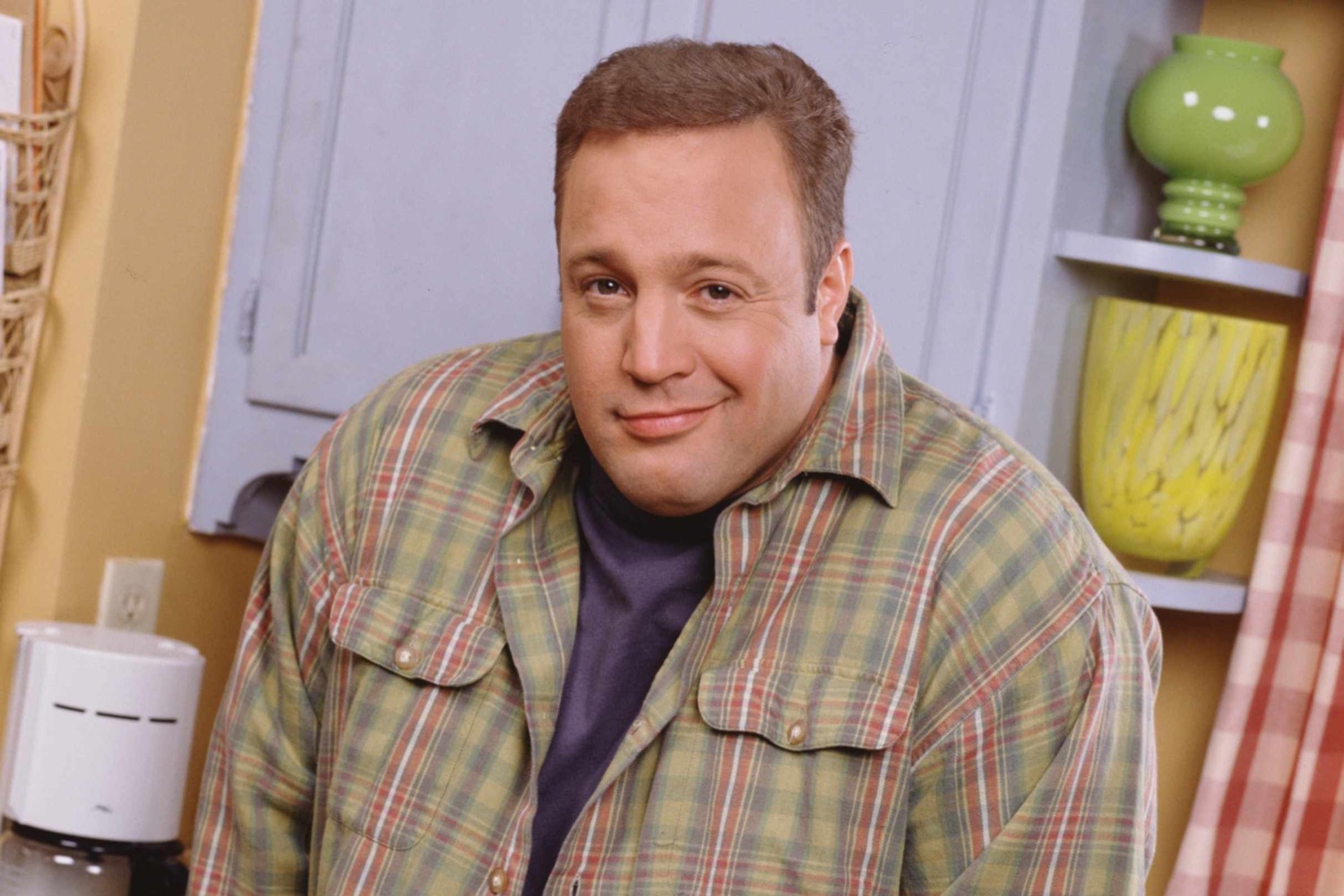 Kevin James in a plaid shirt giving a slightly awkward closed-mouth smirk