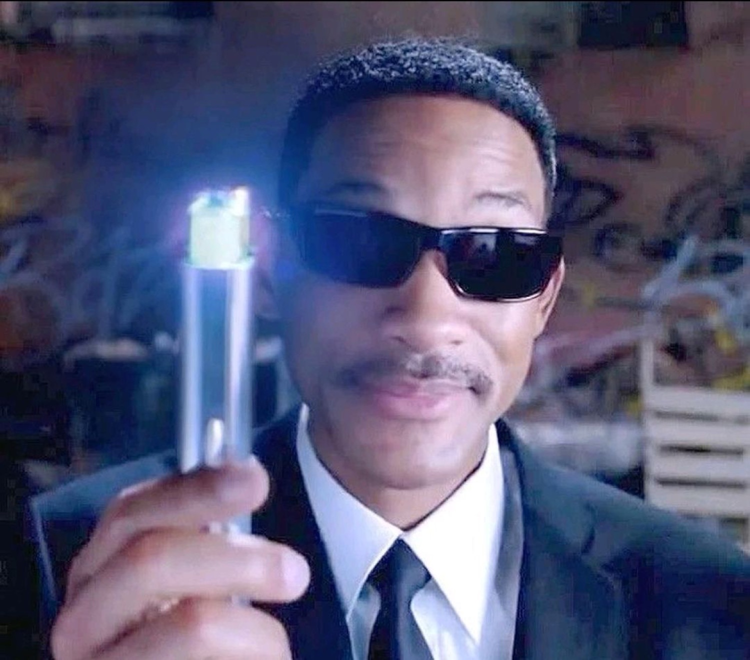 Man in black suit and sunglasses pointing a glowing neuralizer directly at the camera with a smug grin