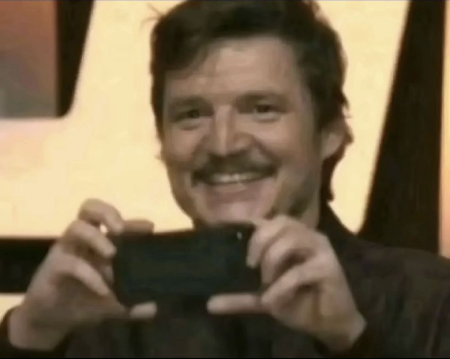 Pedro Pascal smiling while holding up a phone to take a picture