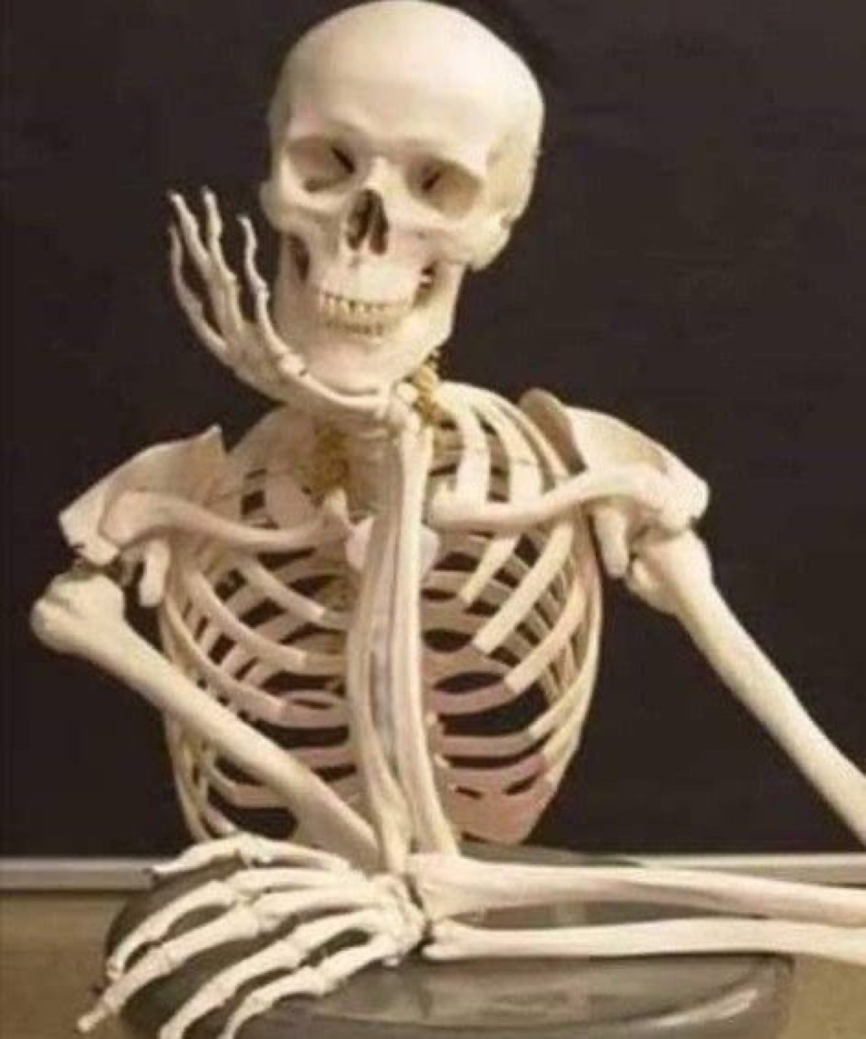 Skeleton sitting at a table with its chin on its hand, waiting forever
