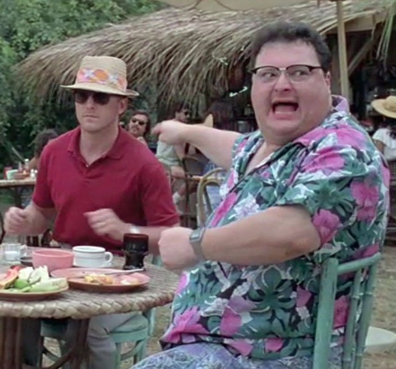Man in a floral shirt mockingly waving his arms at a diner table while a man beside him ignores it