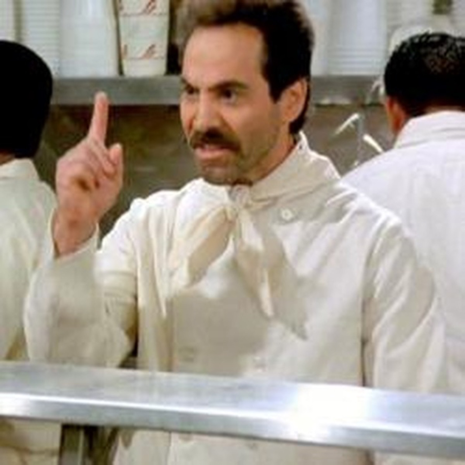 Mustached man in white raising one finger and lecturing sternly behind a counter