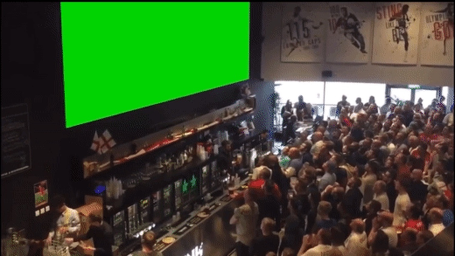 Packed sports bar crowd erupting at a big green screen mid-celebration