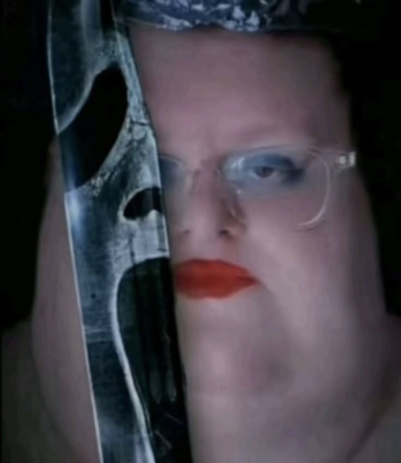 Person in glasses peering around a Ghostface mask with heavy eye makeup and a flat stare
