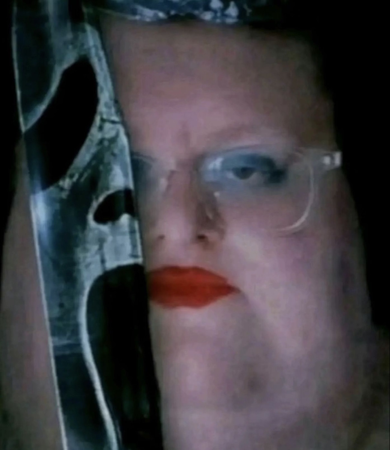 Dim close-up of a face in glasses with red lipstick and a blank, unsettling stare