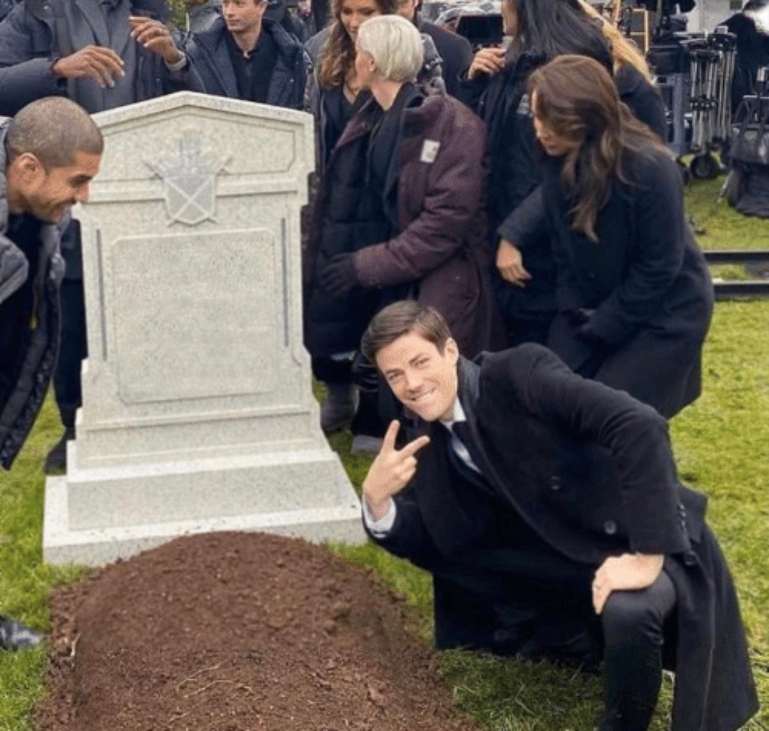 Man in black suit flashing a peace sign and grinning while crouching at a graveside