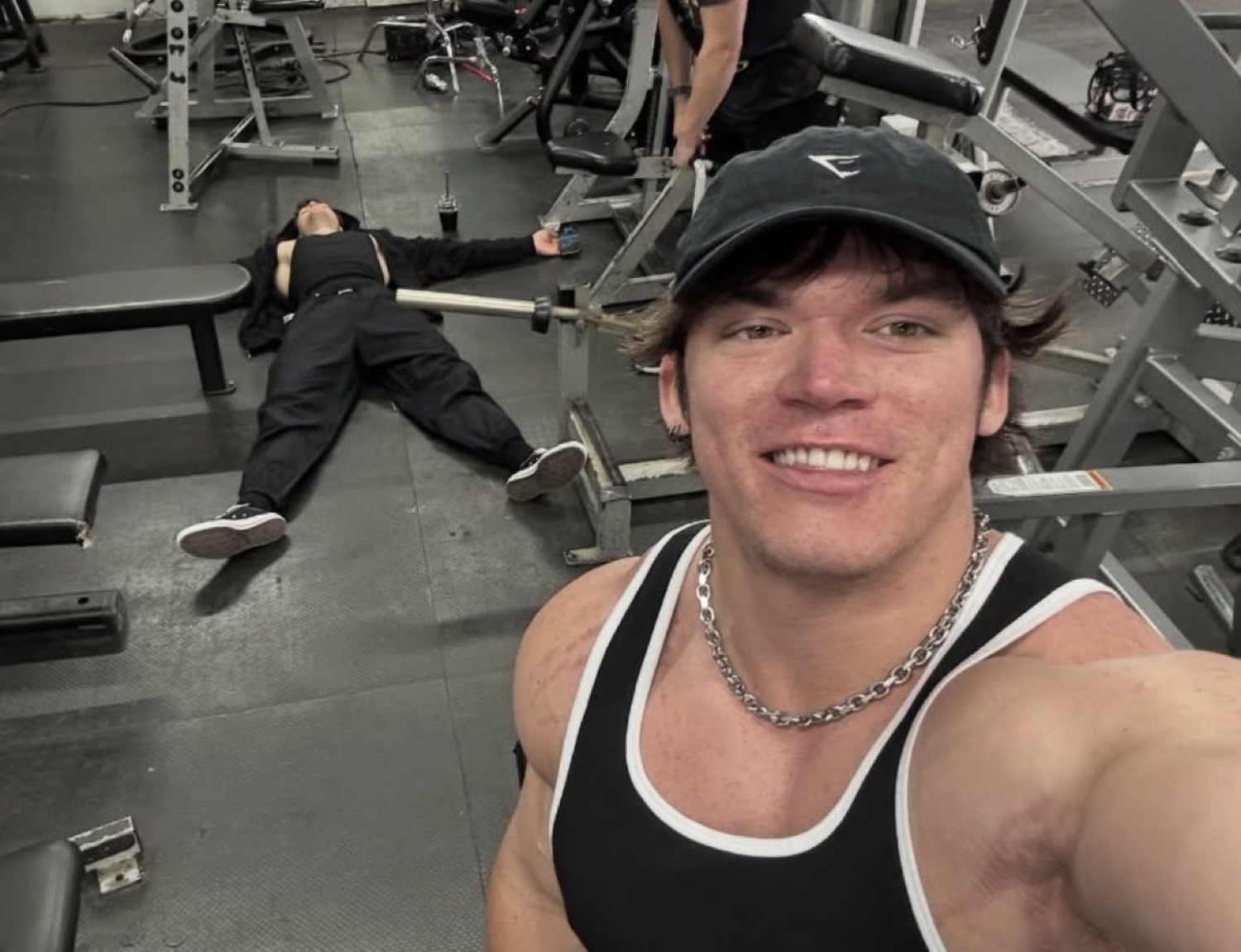 Grinning gym bro taking a selfie while his friend lies collapsed on the floor behind him